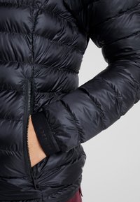 Svart pufferjacka med quiltat mönster, dragkedjefickor framtill och elastiska muddar för en tät passform. Slät, glänsande yttertyg.