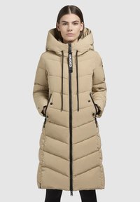 Manteau matelassé beige avec capuche, présentant un design long, une fermeture éclair à l'avant, des poches latérales et un motif en chevrons. Fermeture éclair et cordons noirs.