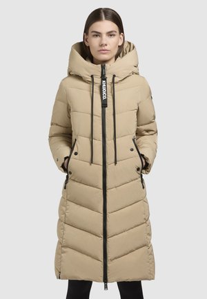 Beige Steppjacke mit Kapuze, lang geschnitten, mit Frontreißverschluss, Seitentaschen und Zickzack-Steppung. Schwarzer Reißverschluss und Kordeln.