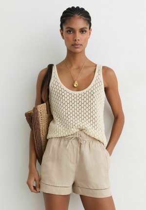 Frau trägt ein cremefarbenes gehäkeltes Tanktop, beige Shorts mit Kordelzug, goldene Anhängerkette, trägt eine gewebte Tragetasche auf der Schulter.
