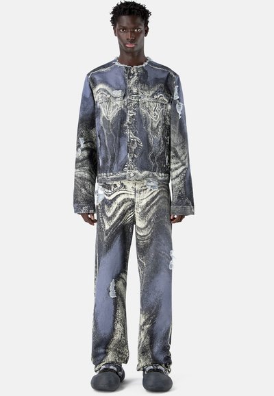 Jeune homme debout portant une veste en denim noir et blanc délavé effet acide et un jean assorti avec des déchirures, ainsi que des chaussures noires sans lacets.