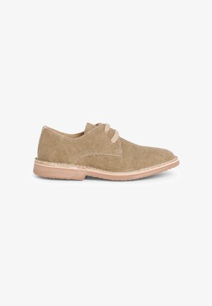 Beige canvas shoe with rounded toe and two lace eyelets. Features a light tan rubber sole with stitching accents along the edges.