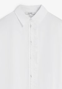 Blusa blanca con un collar clásico, detalles de volantes a lo largo de la botonera y botones de plástico transparente. La tela parece suave y ligera.