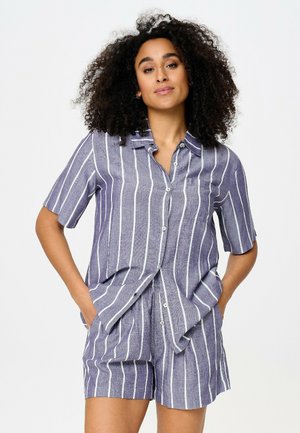 Femme aux cheveux bouclés portant une chemise à boutons à manches courtes rayée verticalement bleu et blanc et un short assorti, les mains dans les poches.