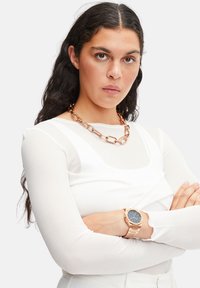 Ur med en mørk urskive, kasse i rosaguld og kædetræk; parret med en hvid langærmet top og et kraftigt halskæde i rosaguld.