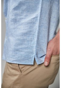 Chemise en lin bleu clair avec un tissage texturé, dotée d'évents latéraux et de détails à boutons, associée à un pantalon kaki.
