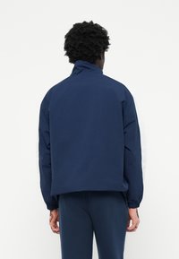 Mørkeblå jakke med lange ærmer, høj krave, elastiske manchetter og hvide detaljer på siderne. Glat materiale, afslappet pasform.