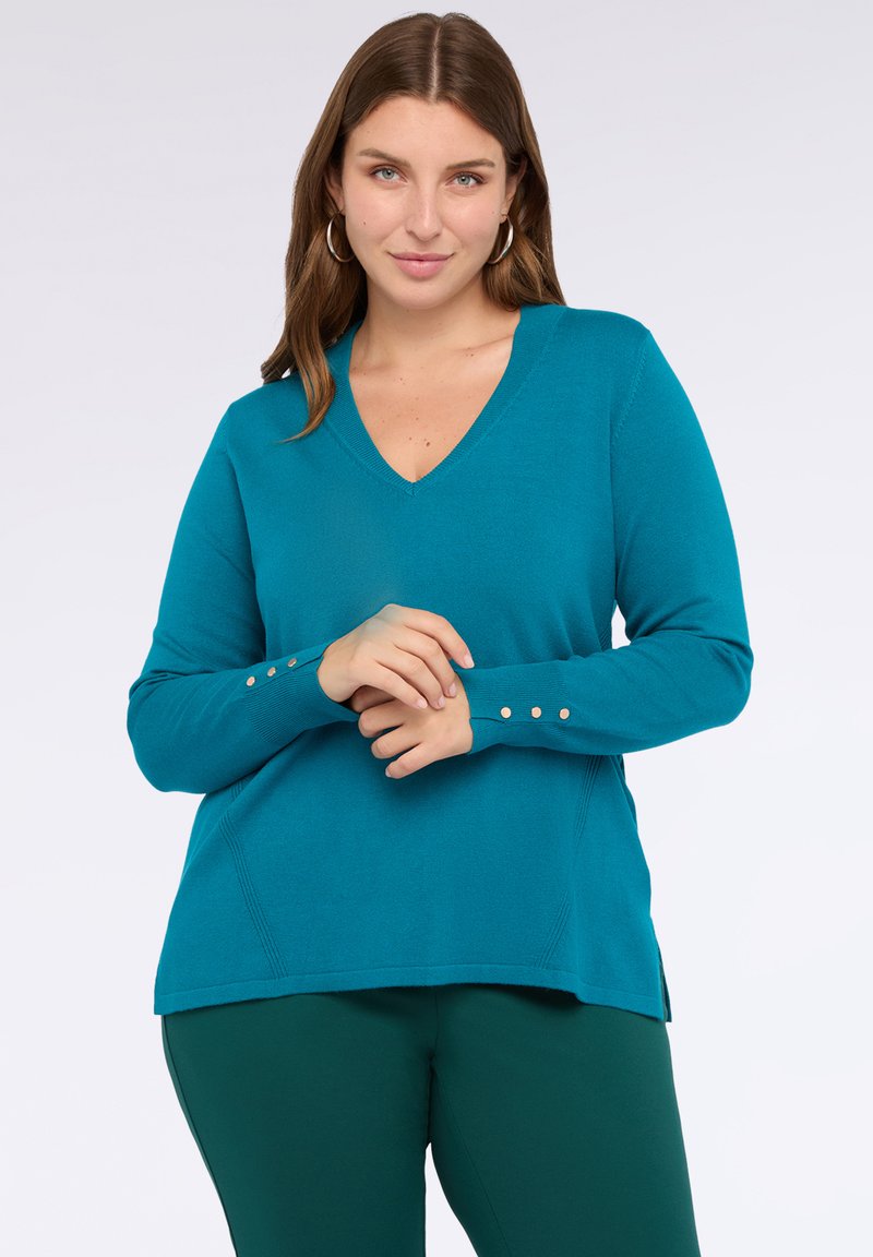 Kobieta w turkusowym swetrze z dekoltem w serek i guzikami na rękawach oraz ciemnozielonych spodniach, pozująca z dłońmi złożonymi na przodzie.