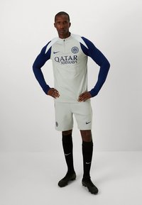 Maillot à manches longues gris clair avec manches bleu marine et logo "Qatar Airways", associé à un short clair et des chaussettes noires, avec un petit logo.