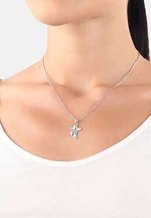 Colgante de ángel de plata con alas sosteniendo un corazón, llevado en el cuello de una persona con una blusa blanca.