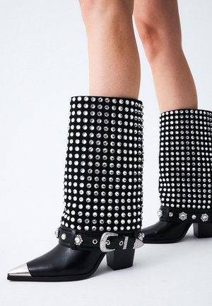 Botas negras hasta la rodilla adornadas con grandes tachuelas plateadas, con una punta metálica puntiaguda y una banda negra con una hebilla decorativa.