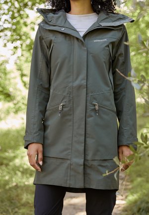 Personne portant un long imperméable vert olive Didriksons avec des poches zippées, debout à l'extérieur parmi un feuillage vert.
