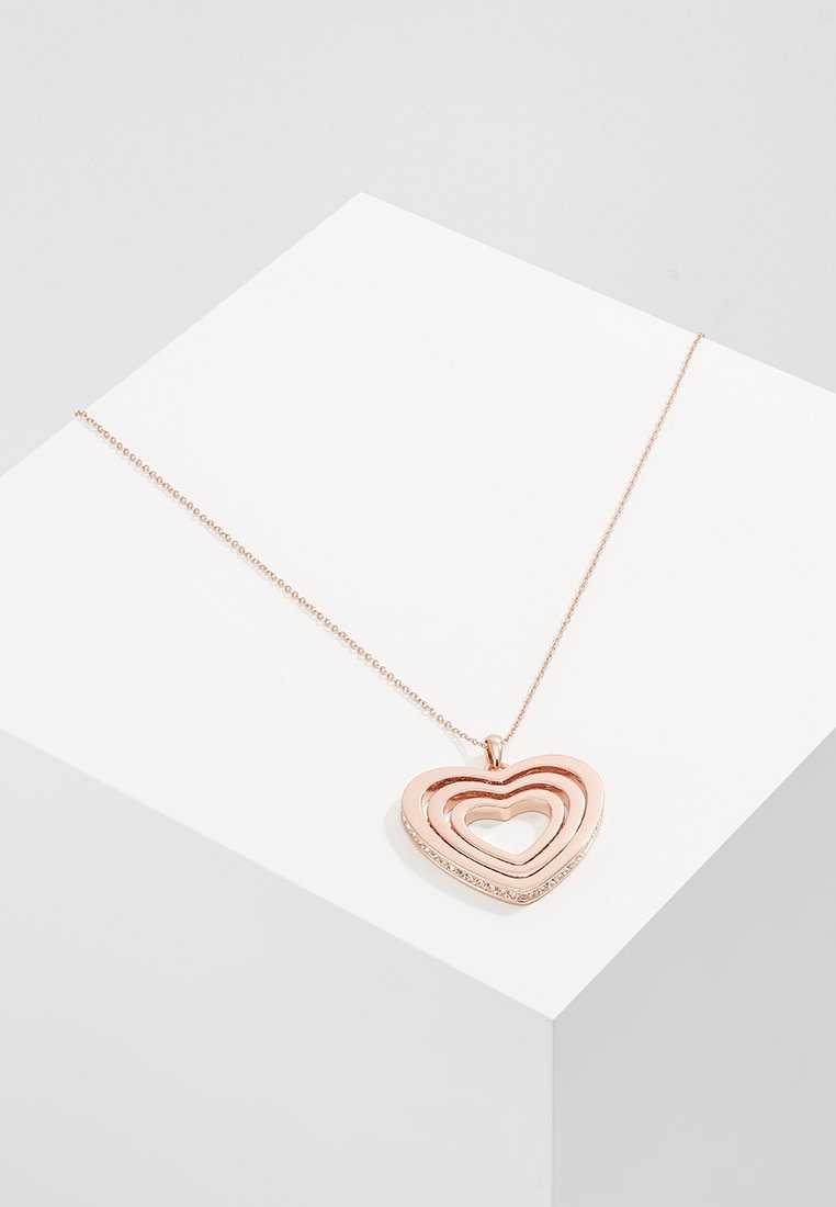 Collier en or rose avec un pendentif présentant trois formes de cœur ouvertes imbriquées, reposant sur une surface géométrique blanche.