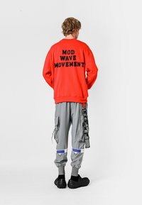 Sudadera roja con "MOD WAVE MOVEMENT" en negro en la parte posterior; combinada con pantalones cargo grises con cremalleras y bolsillos laterales; calzado negro.