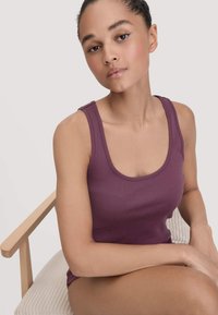 Jonge vrouw met opgestoken haar in een paarse tanktop die op een lichte houten stoel zit tegen een neutrale achtergrond.
