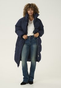 Abrigo inflado de color marino con un cuello alto, cierre frontal con cremallera y botones a presión, combinado con jeans azul ajustados y zapatos negros de punta.