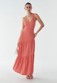 Kobieta ubrana w bezrękawową koralową sukienkę maxi z dekoltem w serek, trzymająca małą białą torebkę, w białych sandałach i złotej biżuterii.