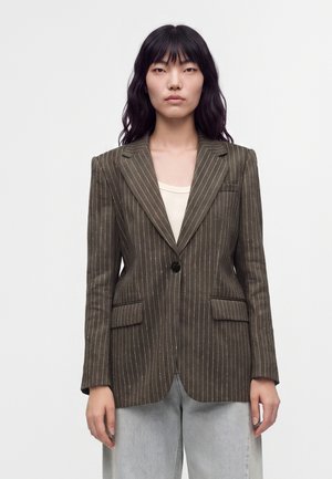 Femme aux longs cheveux noirs porte un blazer marron à fines rayures sur un haut clair et un jean bleu clair, debout devant un fond blanc uni.