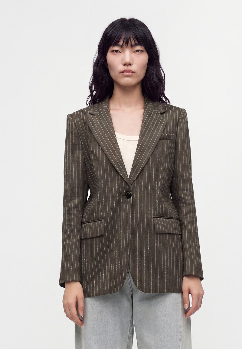 Femme aux longs cheveux noirs porte un blazer marron à fines rayures sur un haut clair et un jean bleu clair, debout devant un fond blanc uni.