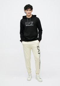 Zwarte katoenen hoodie met "EA7 Emporio Armani"-logo, gecombineerd met crèmekleurige joggingbroek met een logo aan de zijkant en witte sneakers.