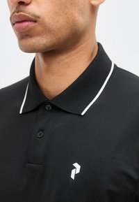 Κοντινό πλάνο ενός ατόμου που φοράει μαύρο πόλο με λευκή διακόσμηση στο γιακά και μικρό λευκό γεωμετρικό λογότυπο στο στήθος.