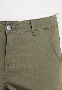 Gros plan sur la taille d'un pantalon vert olive avec un bouton métallique, une passoire de ceinture et une poche latérale, sur un fond de haut blanc.