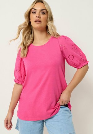 Vrouw met lang blond haar die een felroze top draagt met geborduurde pofmouwen en lichtblauwe denimshorts.