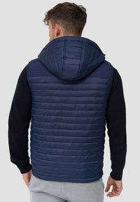 Gilet imbottito blu navy con cappuccio, texture a coste, cuciture orizzontali e maniche nere solide. Design leggero, adatto per essere indossato a strati.