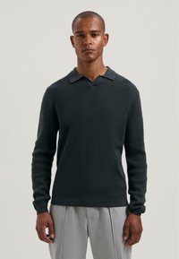Man wearing a dark textured long-sleeve collared sweater and light gray pants, standing against a plain light background.