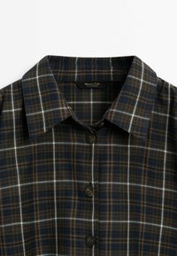 Chemise à carreaux sombre avec des lignes bleues, brunes et blanches, dotée d'un col pointu et de trois boutons noirs sur le devant.