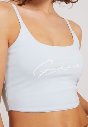 Zbliżenie na kobietę w jasnoniebieskim krótkim topie na ramiączkach z cienkimi paskami i białym logo „Guess” na piersi.