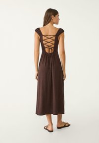 Abito lungo marrone senza maniche con dettaglio in pizzo sulla schiena e spalline a volant. Realizzato in tessuto morbido con una silhouette fluida.