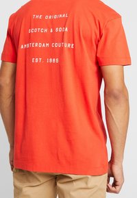 Man in een feloranje T-shirt met witte tekst op de achterkant die zegt "The Original Scotch & Soda Amsterdam Couture Est. 1985."