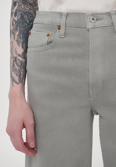 Jean en denim gris à taille haute, avec des accents orange sur les poches, une fermeture à bouton argenté et une finition lisse et texturée.