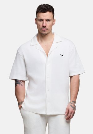 Mann iført hvit kortermet skjorte med knapper og matchende bukser, med tatoveringer på armene og et nøytralt uttrykk, mot ensfarget bakgrunn.