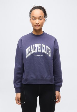 Ung kvinde med krøllet hår iført en marineblå sweatshirt med teksten "HEALTH CLUB BJÖRN BORG", stående foran en ensfarvet lys baggrund.