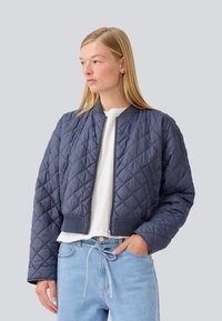 Veste bomber matelassée bleu marine avec fermeture éclair à l'avant, poignets côtelés et ourlet élastique, portée sur une chemise blanche et un jean bleu clair.