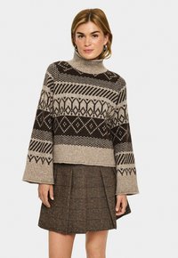 Gestrickter Pullover in Beige und Braun, mit hohem Kragen, weiten Ärmeln und geometrischen Mustern. Kombiniert mit einem braunen Faltenrock.