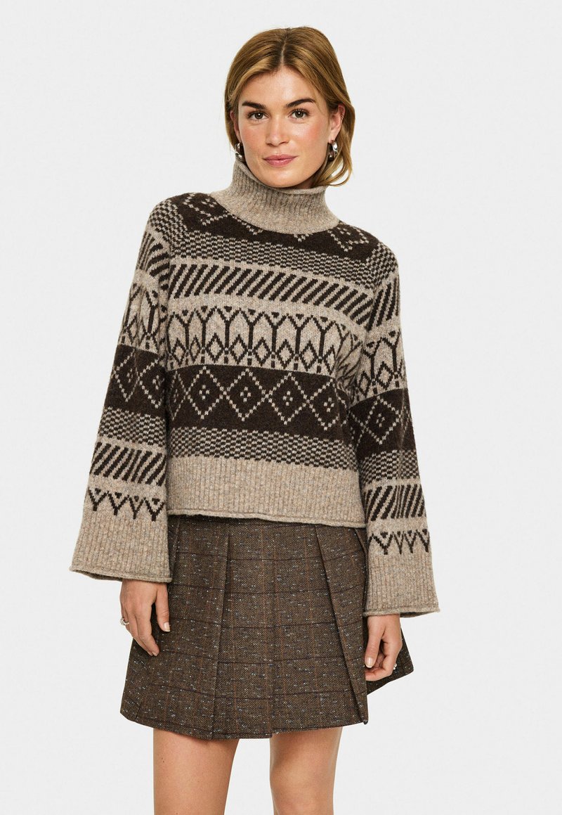 Gestrickter Pullover in Beige und Braun, mit hohem Kragen, weiten Ärmeln und geometrischen Mustern. Kombiniert mit einem braunen Faltenrock.
