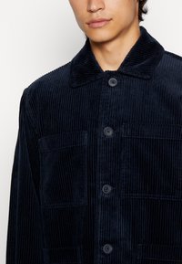 Chaqueta de pana azul marino con cuello, cinco botones frontales y dos bolsillos en el pecho con textura de ribete vertical.