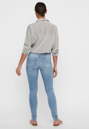 Frau steht mit dem Rücken zur Kamera, trägt ein hellgraues Langarmshirt und hellblaue Skinny-Jeans mit hochgekrempelten Ärmeln sowie rosa Slip-On-Schuhe.