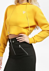 Sudadera amarilla corta con capucha, que presenta cordones plateados y un logo. Combinada con una falda lápiz negra con detalles de cremallera.