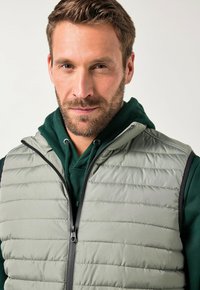 Hellgrüne gep padded Weste mit einem Reißverschluss vorne, ausgestattet mit horizontalen Nähten und einem grünem Kapuzenpullover darunter.