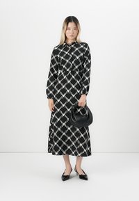 Femme portant une robe à motifs noir et blanc à manches longues, tenant un sac à main noir et portant des escarpins noirs à bouts pointus.