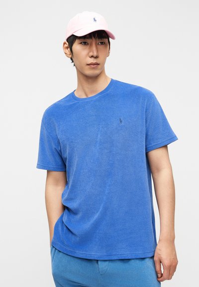 Νέος άνδρας που φοράει μπλε μπλουζάκι από πετσετέ ύφασμα, ταιριαστά μπλε παντελόνια και ένα ανοιχτό ροζ καπέλο μπέιζμπολ, στέκεται μπροστά σε ένα απλό λευκό φόντο.