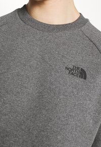 Felpa grigia realizzata in tessuto morbido, con colletto a girocollo a costine, maniche raglan e un logo nero sulla parte sinistra del petto.