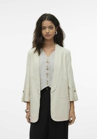 Bež jakk, millel on krae ja mansetid, samuti taskud. Allpool hall, nööbiga särk V-kaelusega. Mustad püksid.