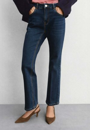 Donna che indossa jeans blu scuro a gamba dritta, tacchi slingback marroni con punta affilata, con le mani nelle tasche su uno sfondo di parete semplice.