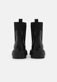 Botines negros con un respaldo texturizado y patrón de cocodrilo. Cuentan con un eje de textil acanalado y una suela de goma gruesa. Dos lengüetas en la parte superior.