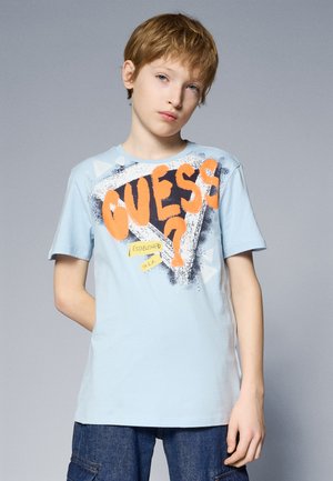 Camiseta de algodón de color azul claro con gran texto naranja "GUESS?" con salpicaduras negras, acentos de etiqueta amarilla y un cuello crew casual.